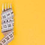 Iron fork and white measuring tape on a yellow background. Diet. Slimming. Obesity. Place for an inscription. Weight loss marathon. Advertising. Top view. The close plan.
